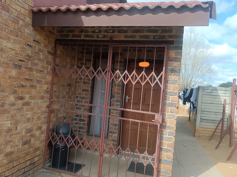 Front entrance of a face brick townhouse featuring a security gate, timber door, and tiled roof overhang.