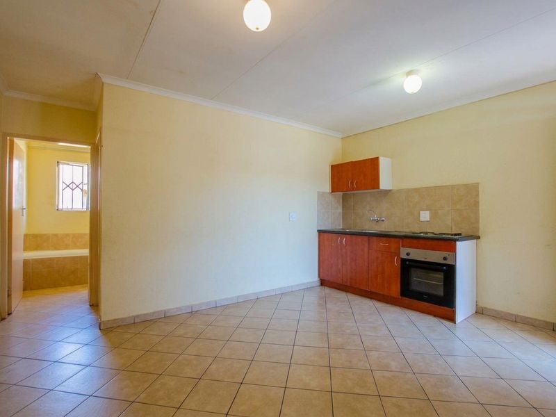 An open-plan living area featuring a compact kitchenette with built-in oven, hob, and cabinetry, with tiled flooring throughout and access to an adjacent bathroom.