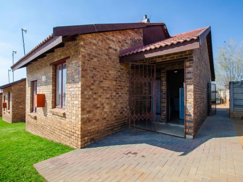 A view of the front exterior of a face brick townhouse featuring a tiled roof, paved entrance area, and a secure metal gate.