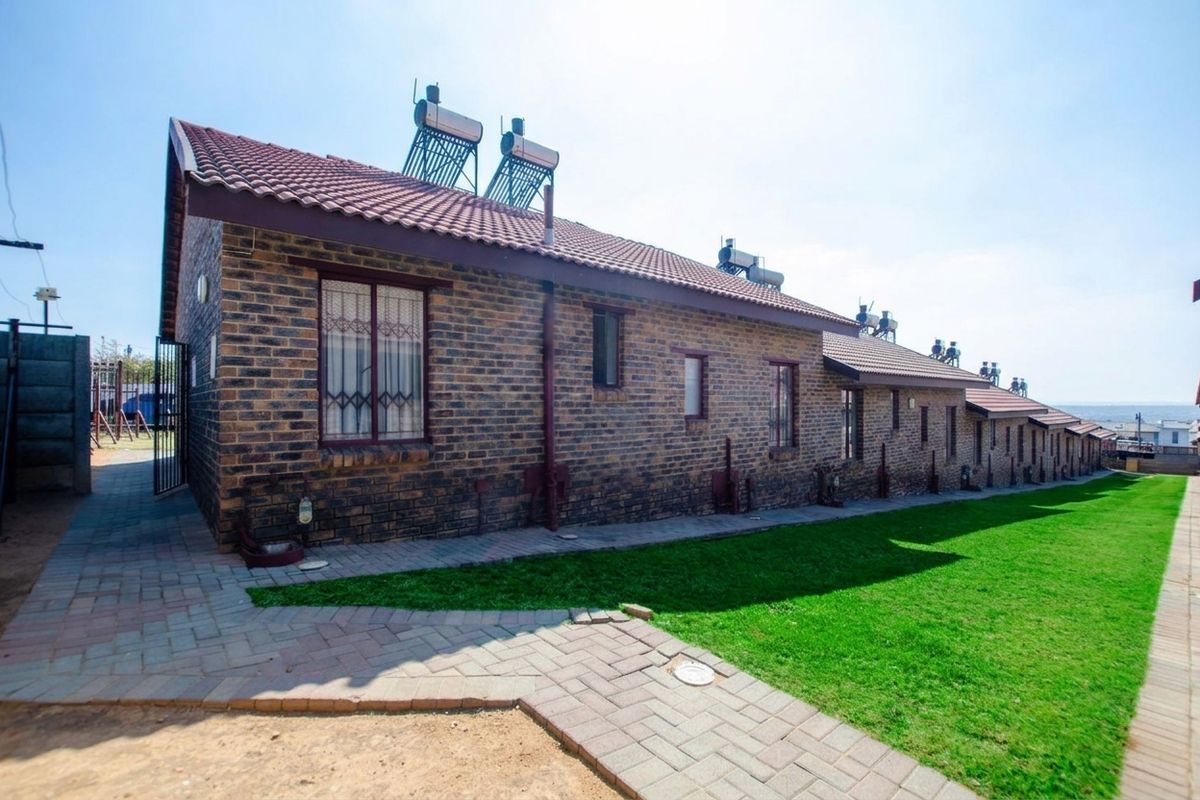 Rear view of a face brick townhouse featuring a tiled roof, solar water heating panels, and a well-maintained lawn with a paved walkway.