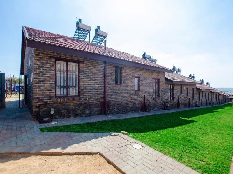 Rear view of a face brick townhouse featuring a tiled roof, solar water heating panels, and a well-maintained lawn with a paved walkway.