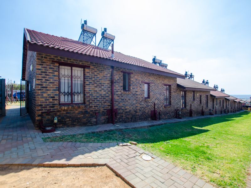 Exterior view of a face brick townhouse row featuring a tiled roof, solar water heating units, and a paved walkway alongside a lawn.
