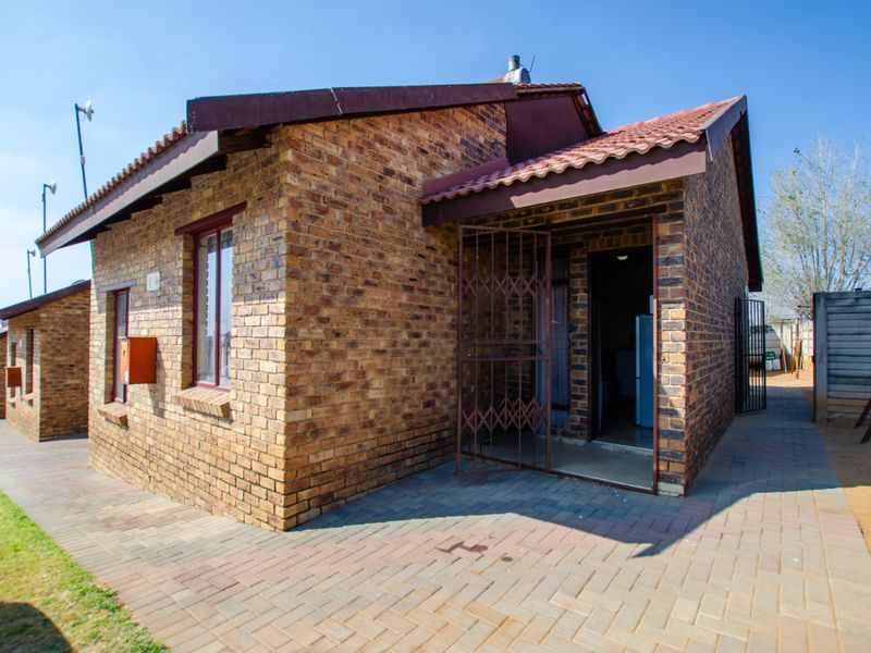 A view of the townhouse exterior featuring face brick construction, a tiled roof, and a paved entrance area with a security gate.