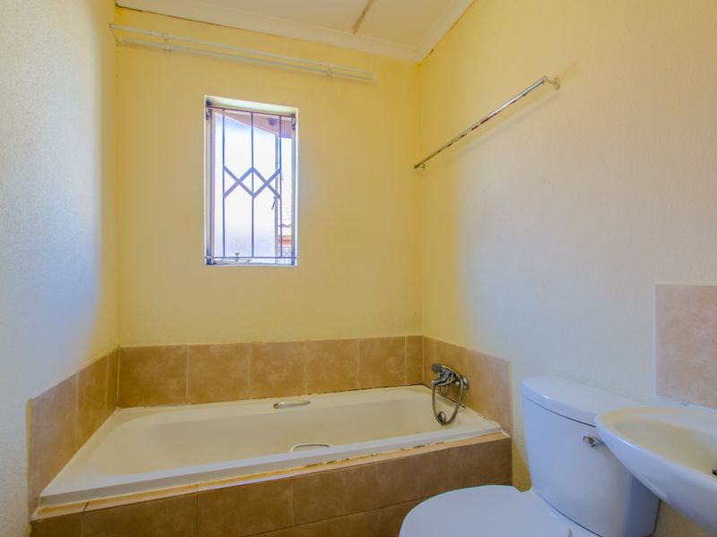A compact bathroom featuring a bathtub, toilet, and washbasin, with security bars on the window and dated wall finishes.