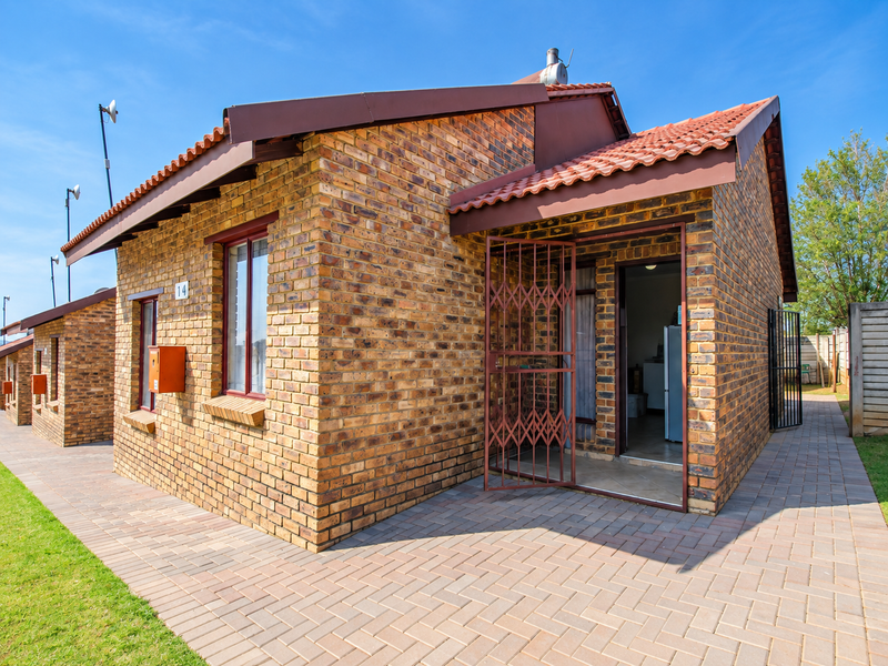 A well-maintained face brick townhouse exterior featuring a tiled roof, secure entrance gate, and paved walkway in a neat residential setting.