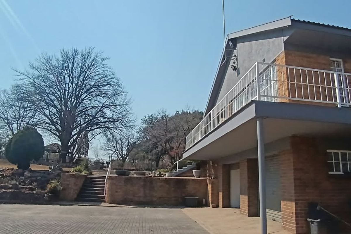 Rear view of a multi-level brick property featuring a balcony with metal railings, paved driveway, and tiered stone landscaping.