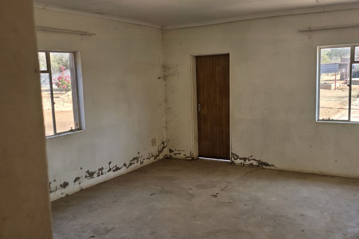 An empty bedroom featuring concrete flooring, steel-framed windows, and a timber door, currently requiring significant maintenance and cosmetic repairs to the walls and ceiling.