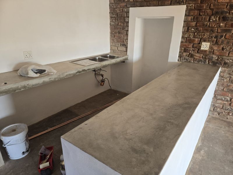 An unfinished kitchen area featuring concrete countertops, a double stainless steel sink, and exposed brickwork, currently undergoing renovation.