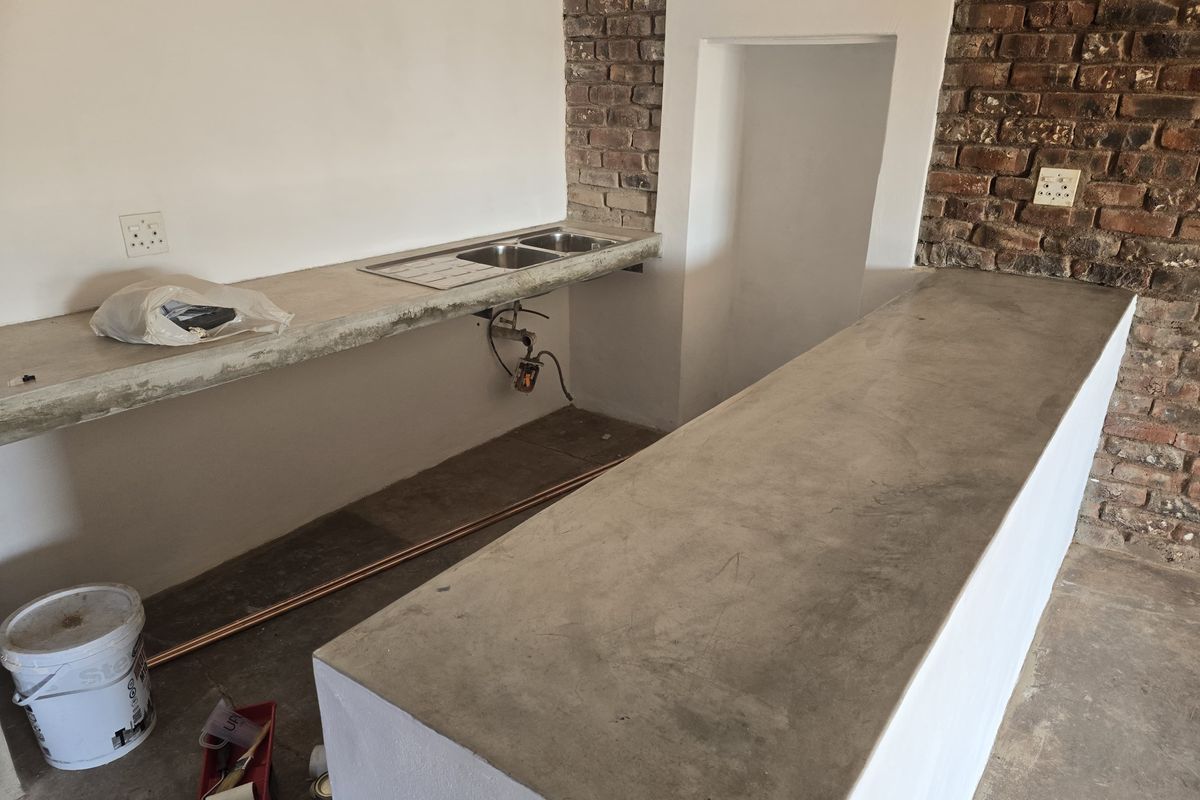 An unfinished kitchen area featuring concrete countertops, a double stainless steel sink, and exposed brickwork, currently undergoing renovation.