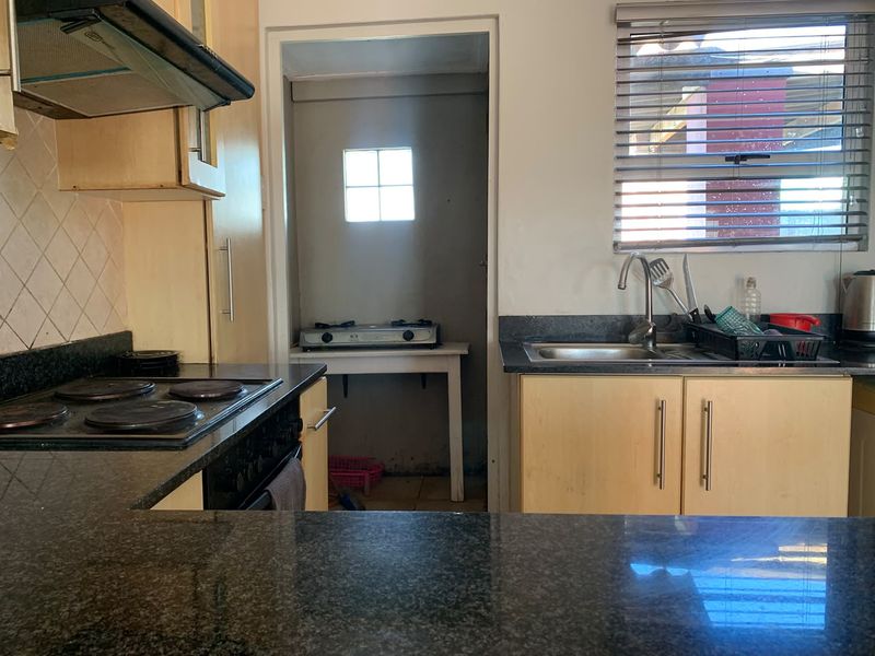 Functional kitchen featuring granite countertops, built-in cabinetry, electric hob, extractor fan, and a stainless steel sink with a view through venetian blinds.