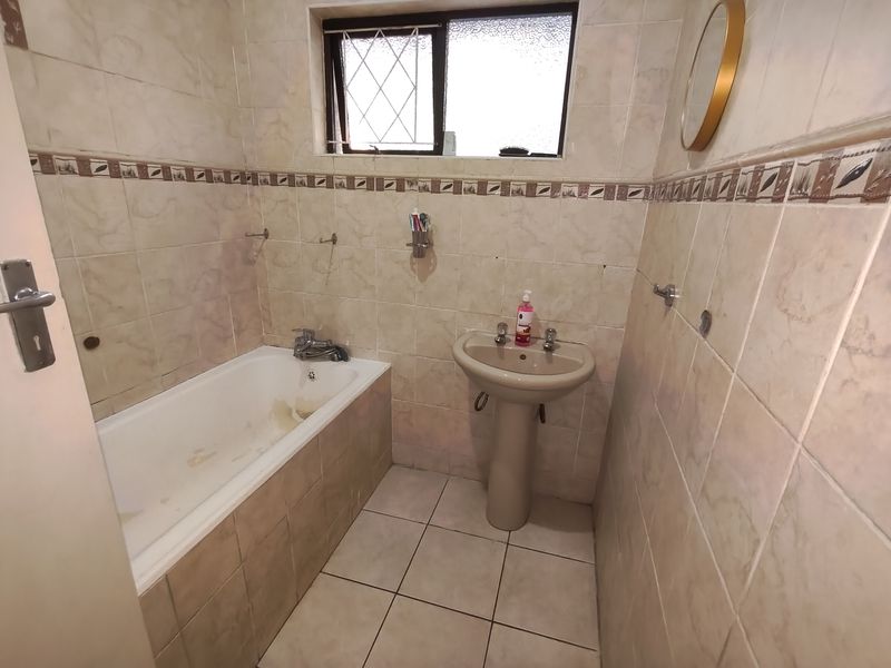 A dated bathroom featuring a built-in bathtub, pedestal sink, and full-height ceramic wall tiling, requiring cosmetic updates to modernize the space.