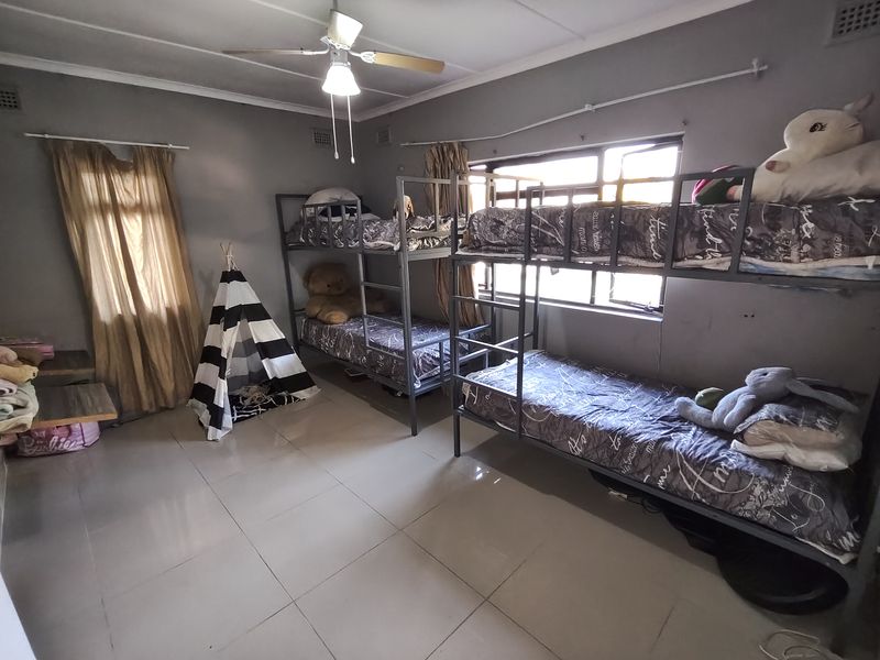 Spacious bedroom featuring tiled flooring, a ceiling fan, and large windows, providing a functional layout suitable for multiple beds.