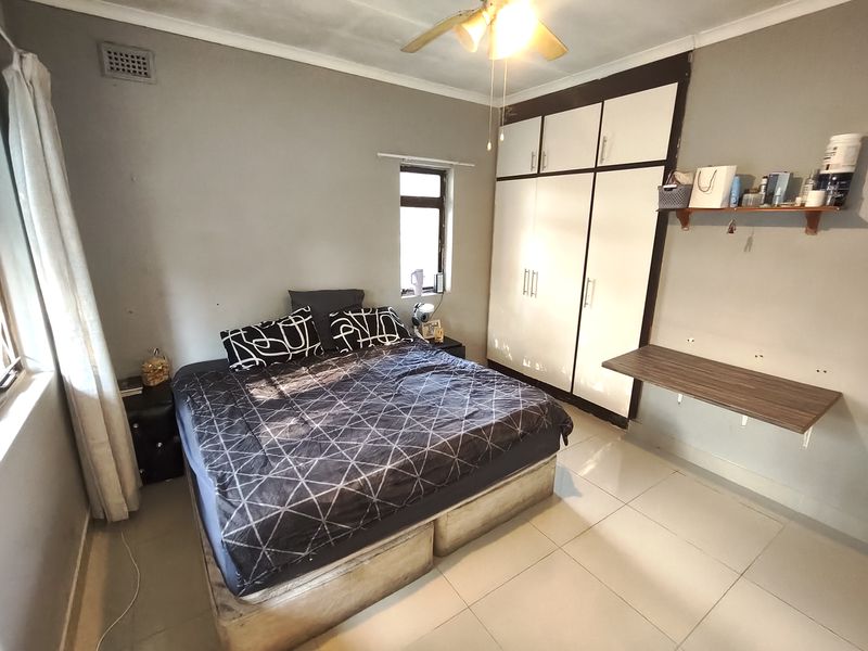 A functional bedroom featuring built-in cupboards, a ceiling fan, and tiled flooring, offering a practical layout with a wall-mounted desk and shelving.