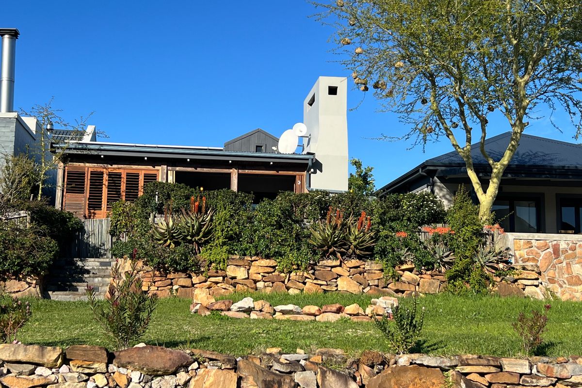 Rear view of the property featuring a landscaped garden with stone retaining walls, mature trees, and a covered patio area with timber shutters.