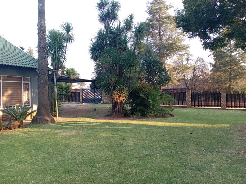 A spacious, well-maintained garden featuring a lush lawn, mature trees, and secure perimeter fencing with a metal driveway gate.
