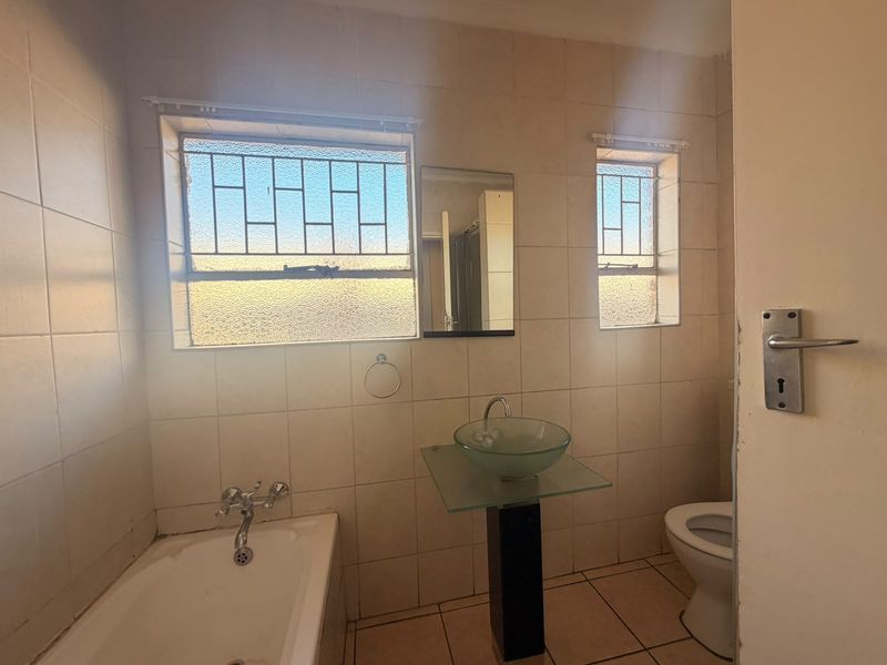 A dated bathroom featuring a bathtub, pedestal basin, and toilet, with full-height wall tiling and security bars on the windows.