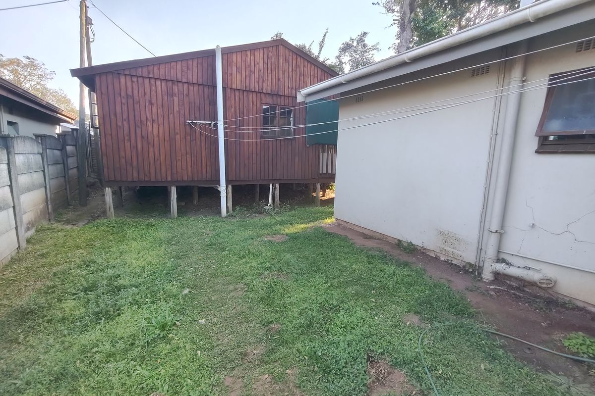 Rear exterior view showing a timber-clad structure on stilts adjacent to a main dwelling with a small grassed yard and boundary wall.