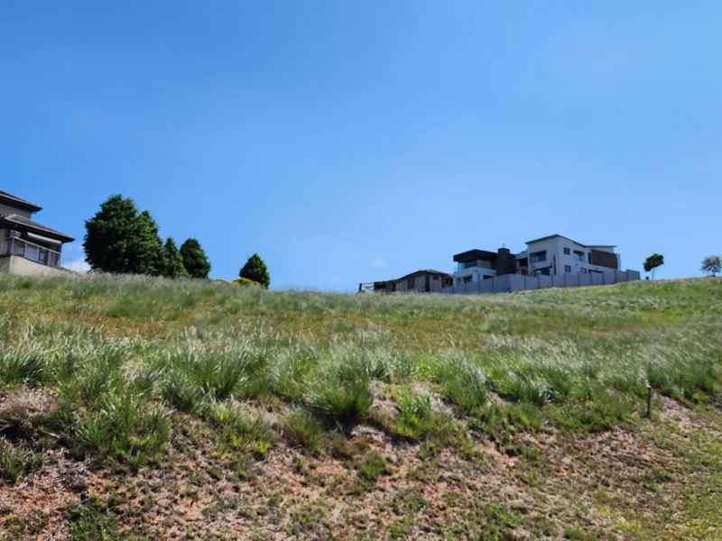 A view of a vacant residential plot featuring a gentle slope, natural grass cover, and an adjacent paved driveway in a developed neighborhood.