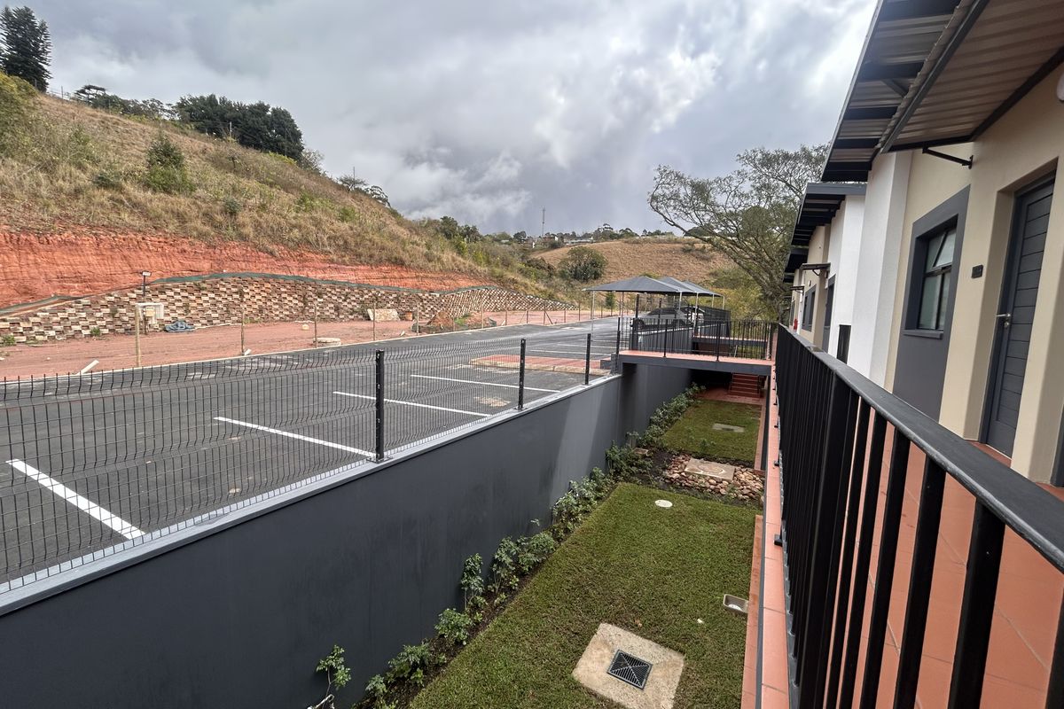 View from the apartment balcony overlooking a landscaped garden, paved parking area, and surrounding hillside.