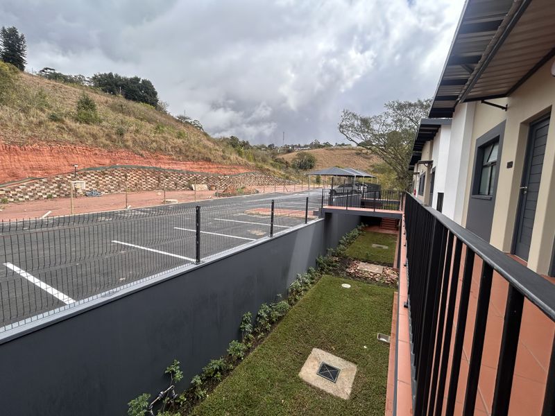 View from the apartment balcony overlooking a landscaped garden, paved parking area, and surrounding hillside.