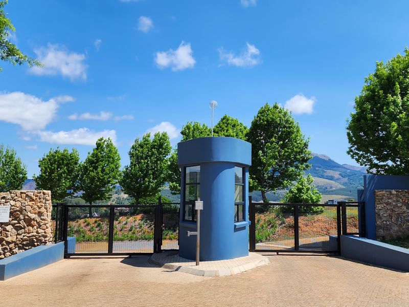 A secure gated entrance featuring a blue guard house, stone masonry walls, and a paved driveway leading into the residential estate.
