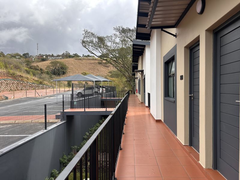A clean, covered exterior balcony walkway featuring tiled flooring and metal balustrades, providing access to apartment units with views of the parking area.