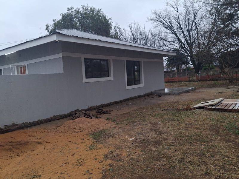 Rear view of a single-story house featuring a corrugated metal roof, aluminium window frames, and an unfinished outdoor area.