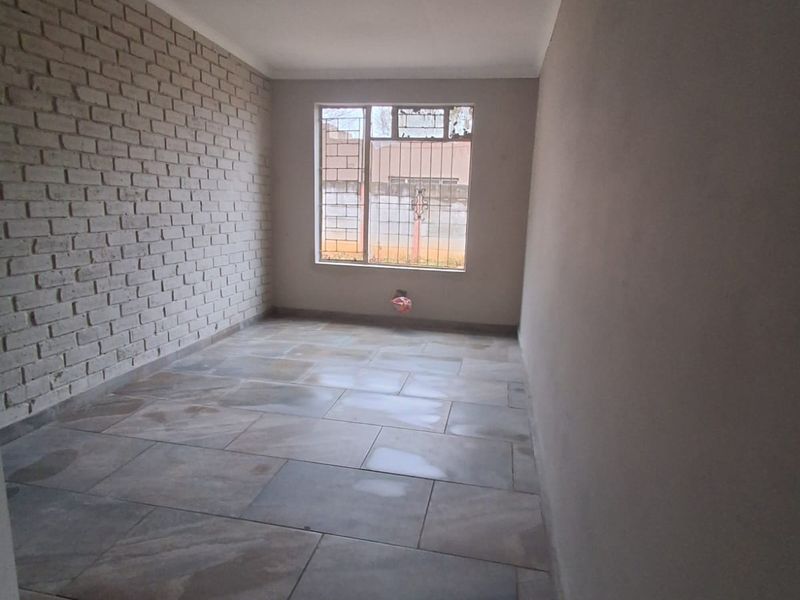 An empty living room featuring a textured exposed brick wall, large format floor tiles, and a window with security bars.