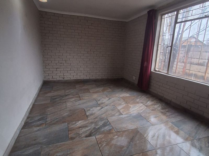 An empty living room featuring exposed brick walls, large windows with security bars, and durable tiled flooring.