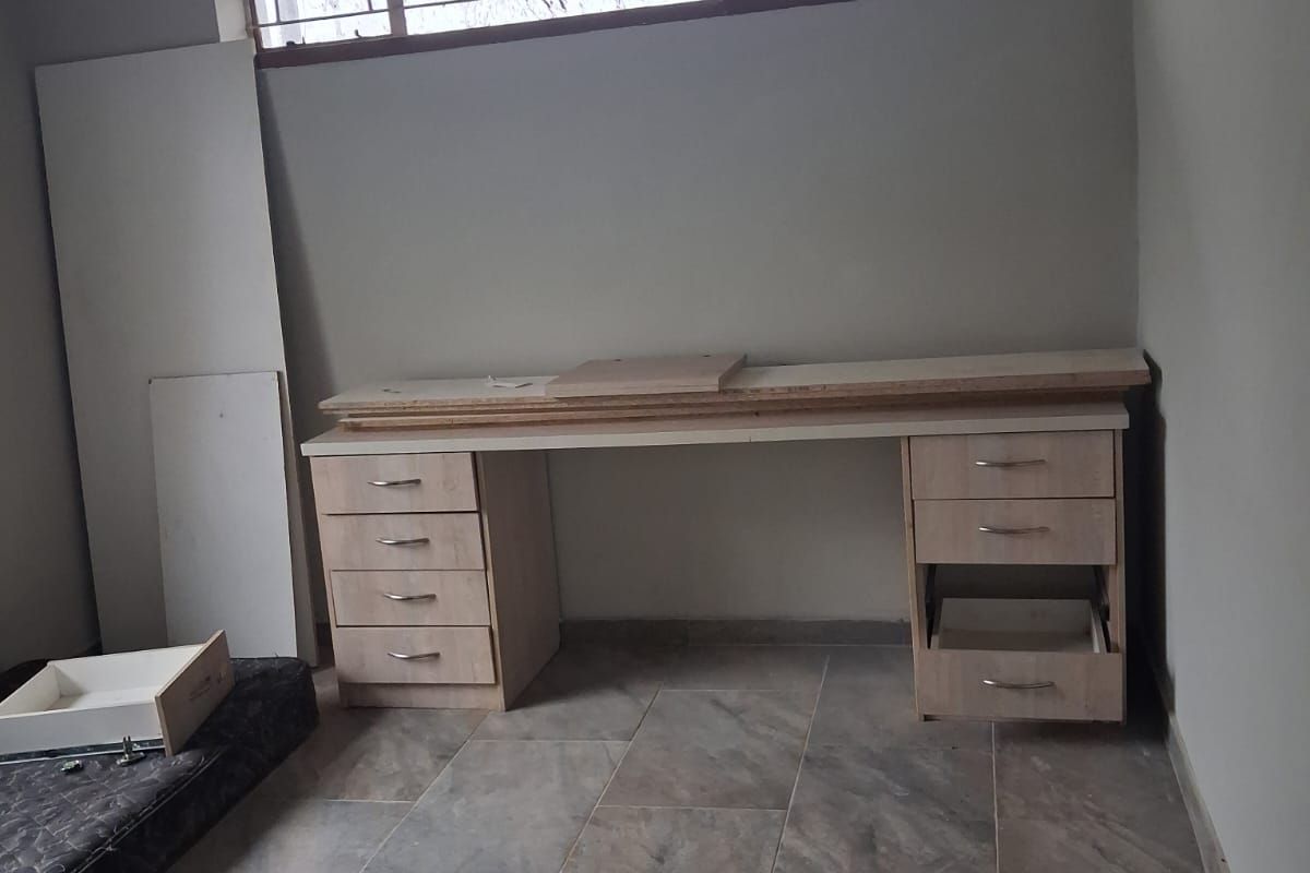A bedroom featuring tiled flooring, a high-set window with security bars, and a built-in desk unit requiring completion and repair.