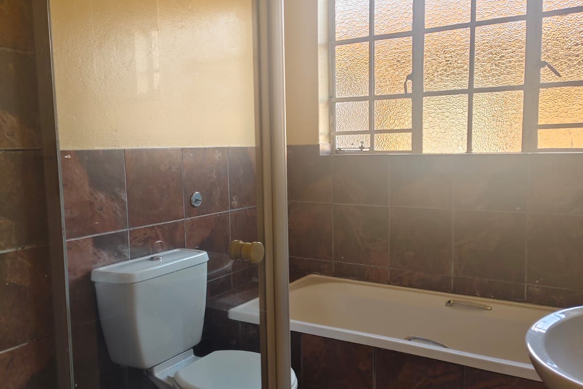 A functional bathroom featuring a built-in bathtub, toilet, and pedestal basin with dark tiled finishes and a grid-style window.