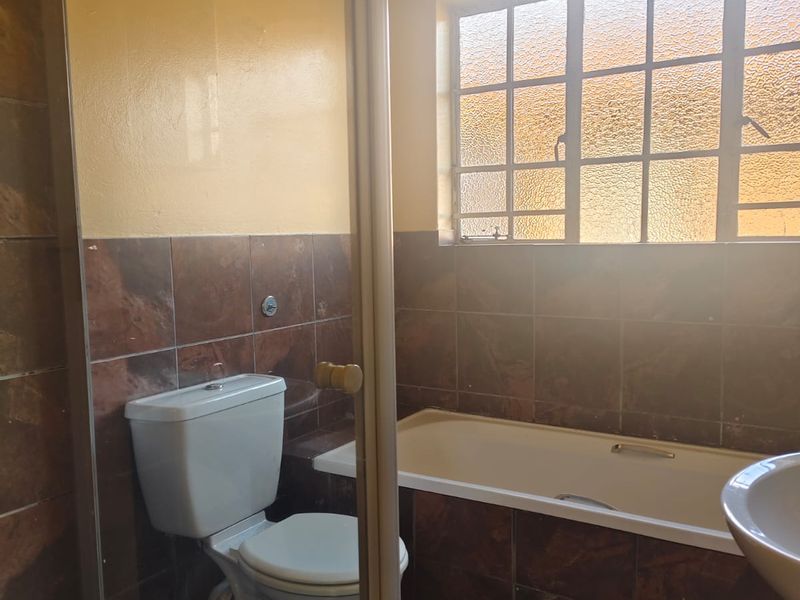 A functional bathroom featuring a built-in bathtub, toilet, and pedestal basin with dark tiled finishes and a grid-style window.