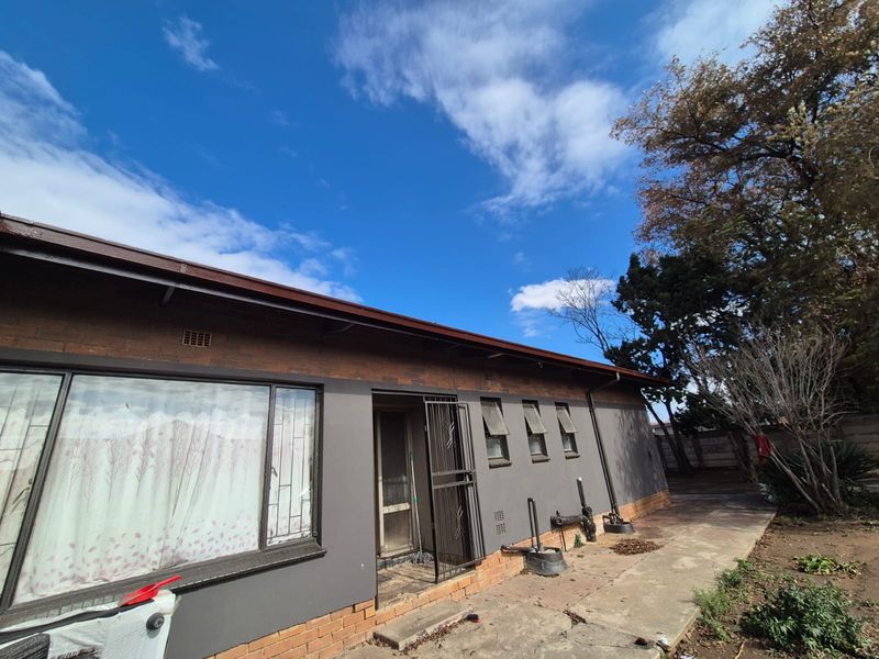 Rear exterior view of a brick house featuring a concrete pathway, security gate, and multiple casement windows under a clear blue sky.