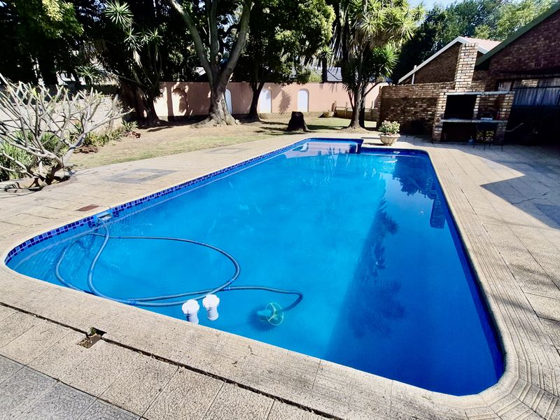 Outdoor entertainment area featuring a swimming pool, paved patio, and a built-in brick braai, set within a private garden with mature trees.
