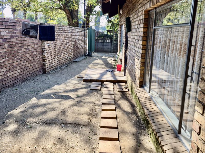 A side or rear exterior area featuring a brick boundary wall, paved walkway, and large windows, offering potential for landscaping and outdoor improvement.