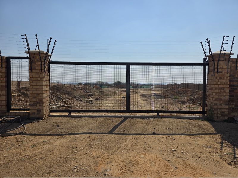 A secure entrance featuring brick pillars with electric fencing and a large metal sliding gate, providing access to the vacant residential plot.