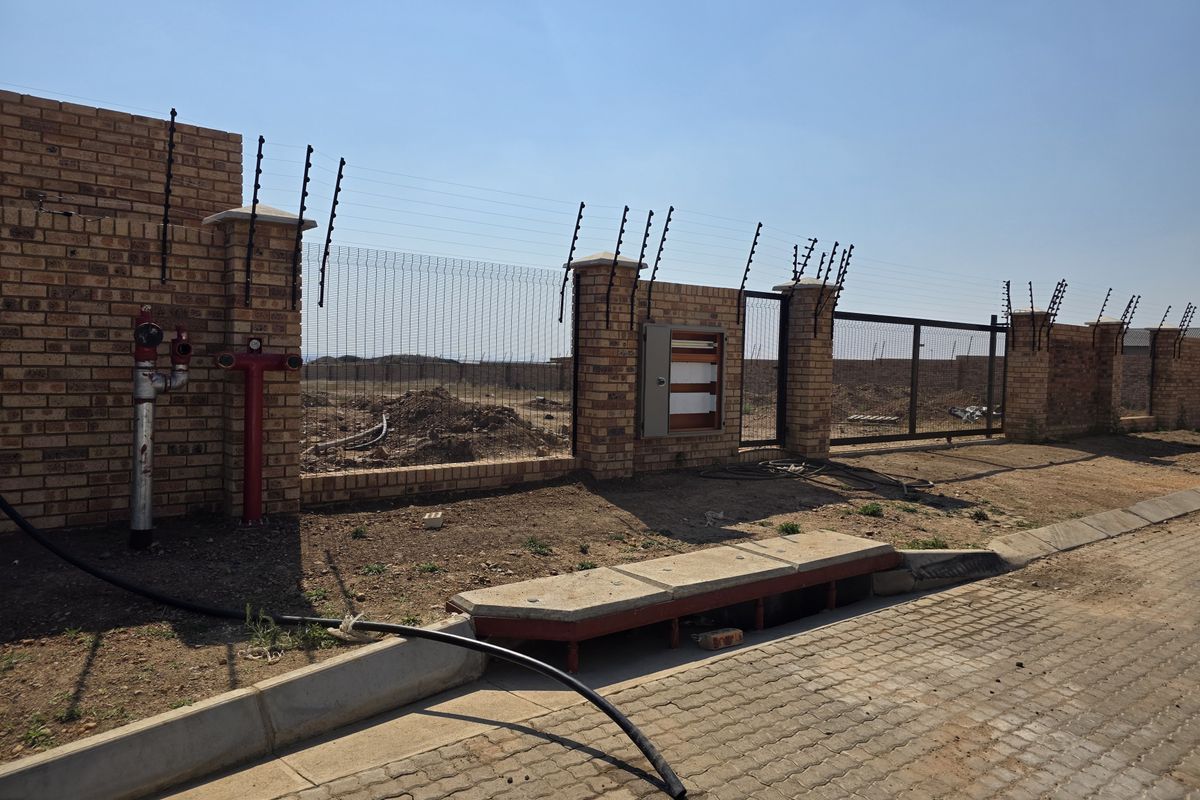 Front view of a vacant residential plot featuring a brick boundary wall, electric fencing, fire hydrant, and utility access point.