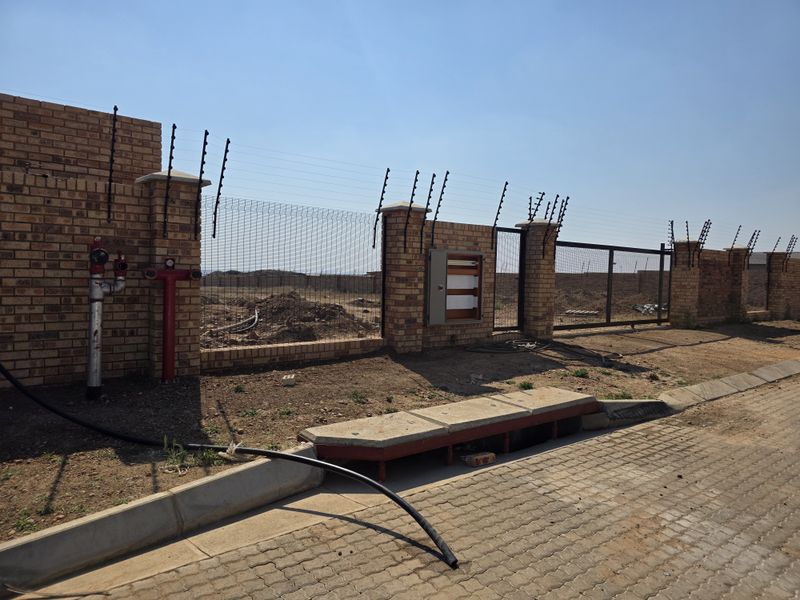 Front view of a vacant residential plot featuring a brick boundary wall, electric fencing, fire hydrant, and utility access point.