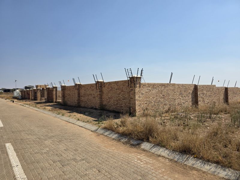 A vacant residential plot enclosed by a secure face brick perimeter wall with installed electric fencing, situated along a paved street.