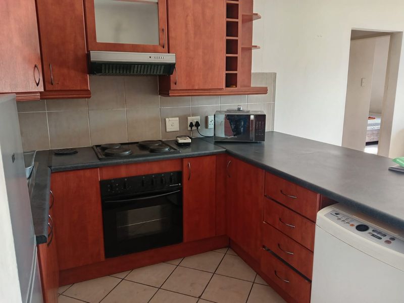 A functional kitchen featuring wooden cabinetry, dark laminate countertops, an integrated electric hob, oven, and extractor fan with tiled flooring and backsplash.