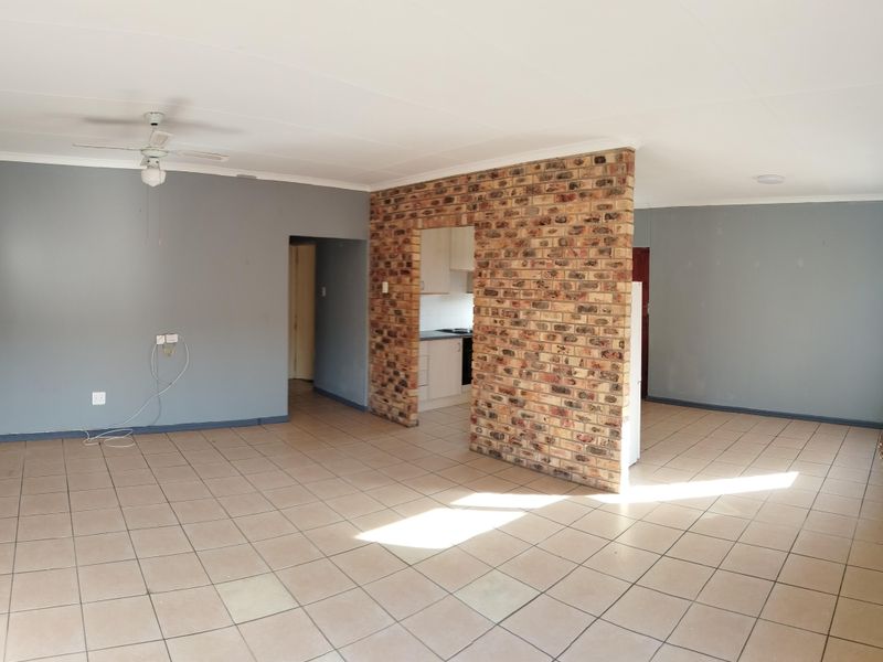 Spacious open-plan living area featuring a distinctive exposed brick wall, neutral tiled flooring, and a ceiling fan, offering a versatile layout for modern living.