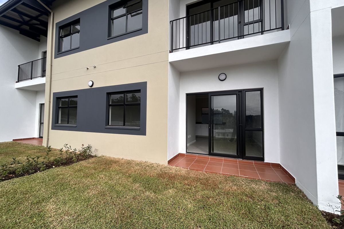 Rear view of a modern apartment building featuring a ground-floor tiled patio, sliding glass doors, and upper-level balconies with metal railings.