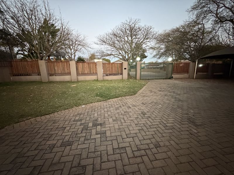 Front exterior view featuring a paved driveway, manicured lawn, and secure perimeter wall with wooden fencing and a metal gate.