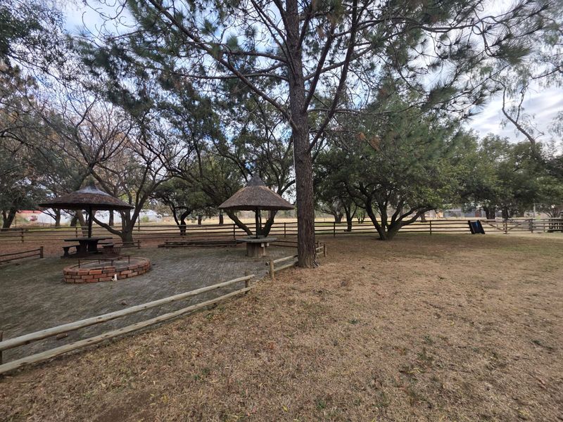 Spacious garden area featuring two thatched lapas, a brick fire pit, paved patio space, and mature trees enclosed by timber fencing.