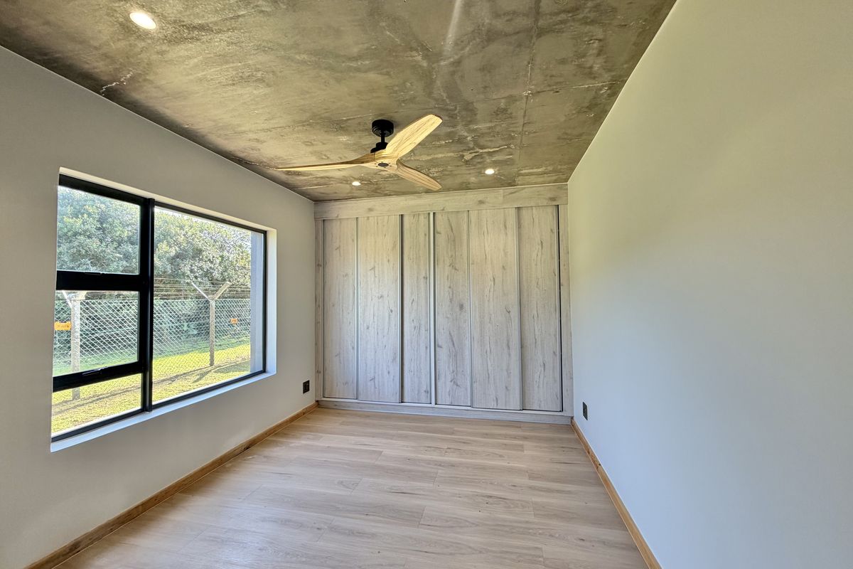 A modern, empty bedroom featuring an exposed concrete ceiling, light wood-look laminate flooring, and built-in sliding door cupboards.