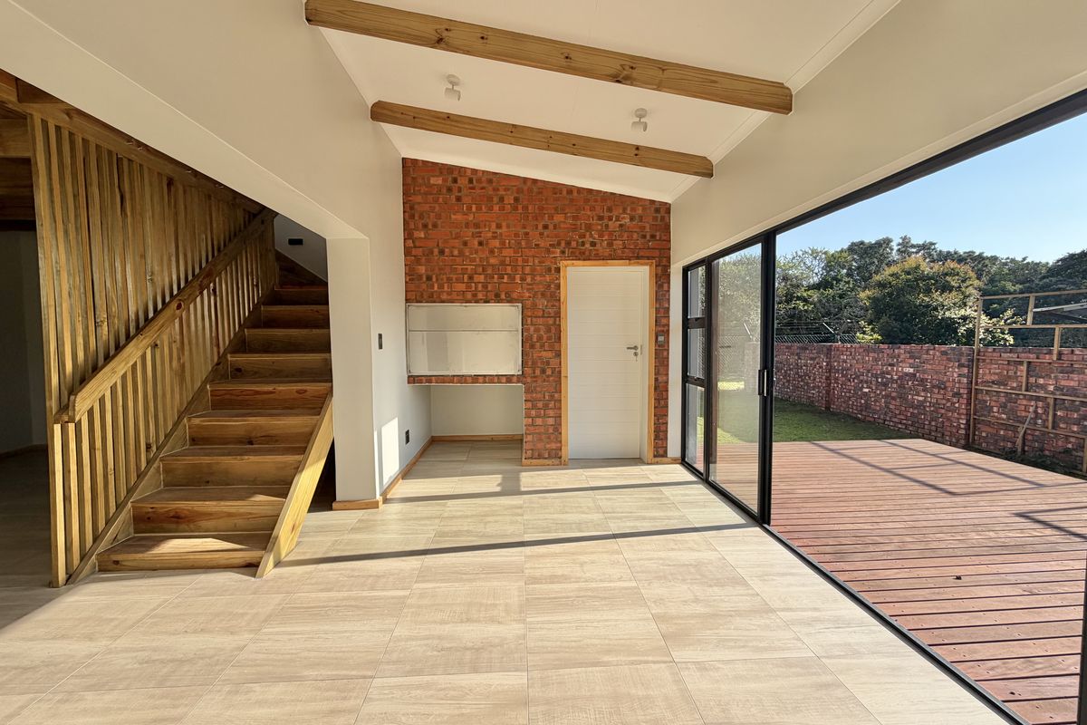 Spacious open-plan living area featuring exposed brickwork, timber ceiling beams, a built-in braai, and large sliding doors opening onto a wooden deck.