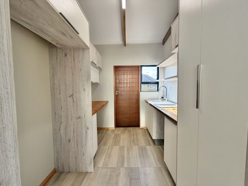 A clean, modern scullery featuring light wood-grain cabinetry, timber countertops, a stainless steel sink, and neutral tiled flooring.