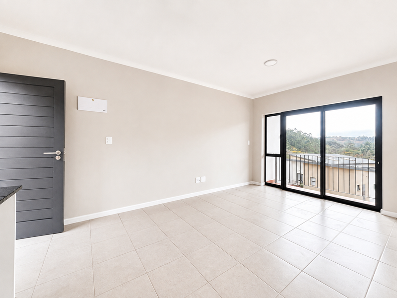 A bright, empty living area featuring neutral tiled flooring, a modern dark-toned door, and large sliding glass doors leading to a balcony with scenic views.