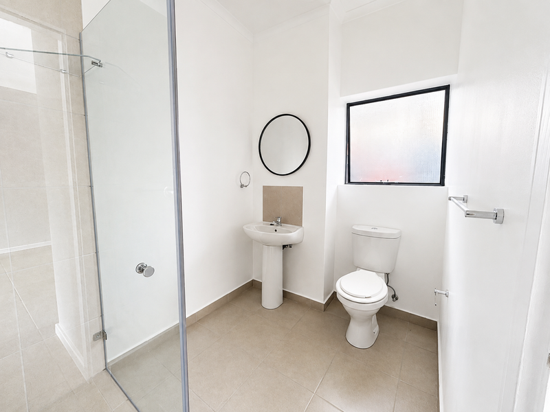 A clean, modern bathroom featuring a pedestal basin, toilet, and a glass-enclosed shower with neutral floor tiling and natural light.