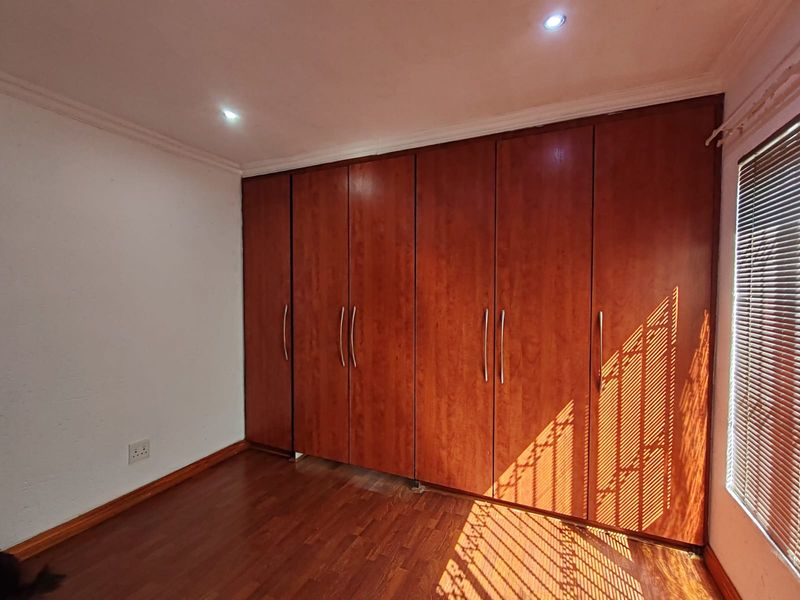 A clean bedroom featuring extensive built-in wooden cupboards, warm laminate flooring, and venetian blinds, offering a functional and well-maintained storage solution.